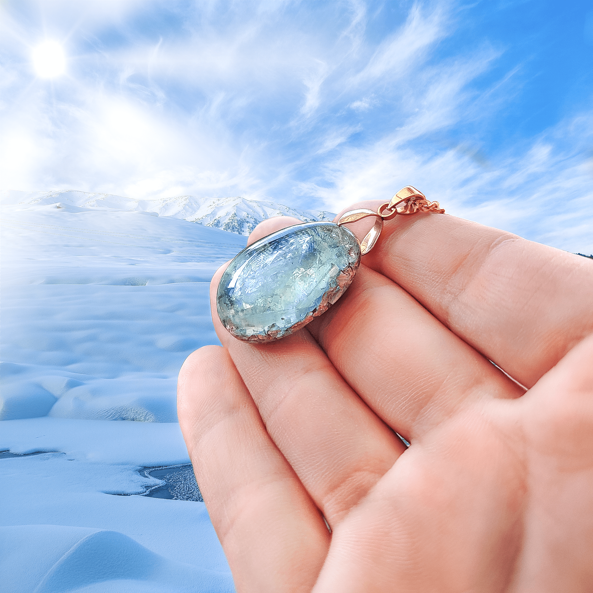 Ein eisblauer Orgonit Anhänger aus Edelsteinen, welcher in einer Schneebedeckten Umgebung ins Licht gehalten wird. Befestigt ist das Amulett an einer vergoldeten Kette.