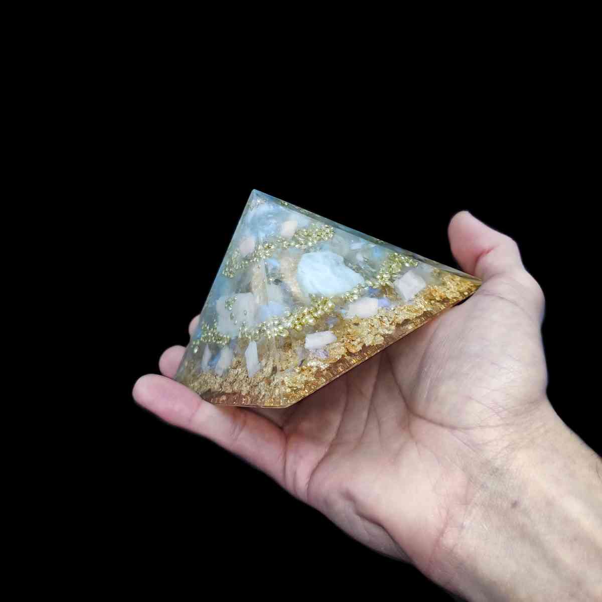 Orgonit Pyramide mit Regenbogenmondstein & Gold, welche von einer Hand gehalten wird.