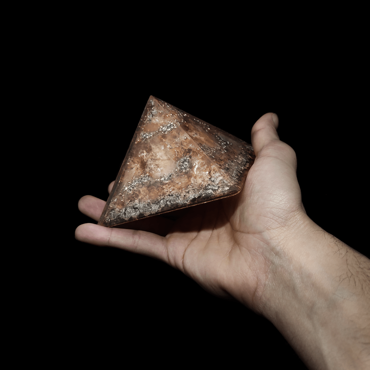 Hellbrauner Orgonit Pyramiden Kristall mit silbernen Elementen, welcher von einer Hand gehalten wird.