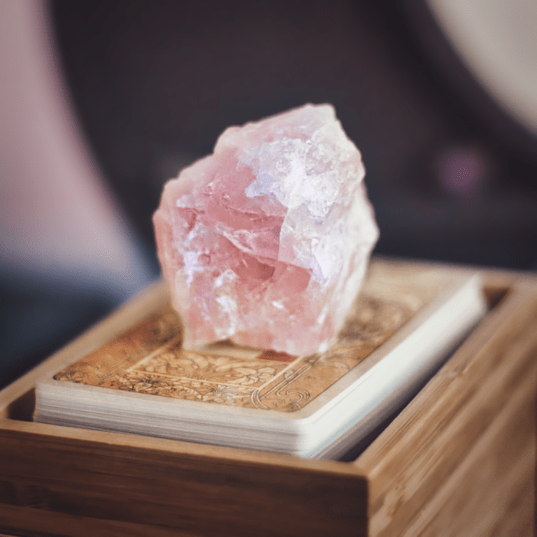 Ein farbintensiver Rosenquarz Rohstein. Dieser Kristall steht auf einem Buch & dient als Symbolbild für unseren Blogbeitrag über die Bedeutung von Rosenquarz.
